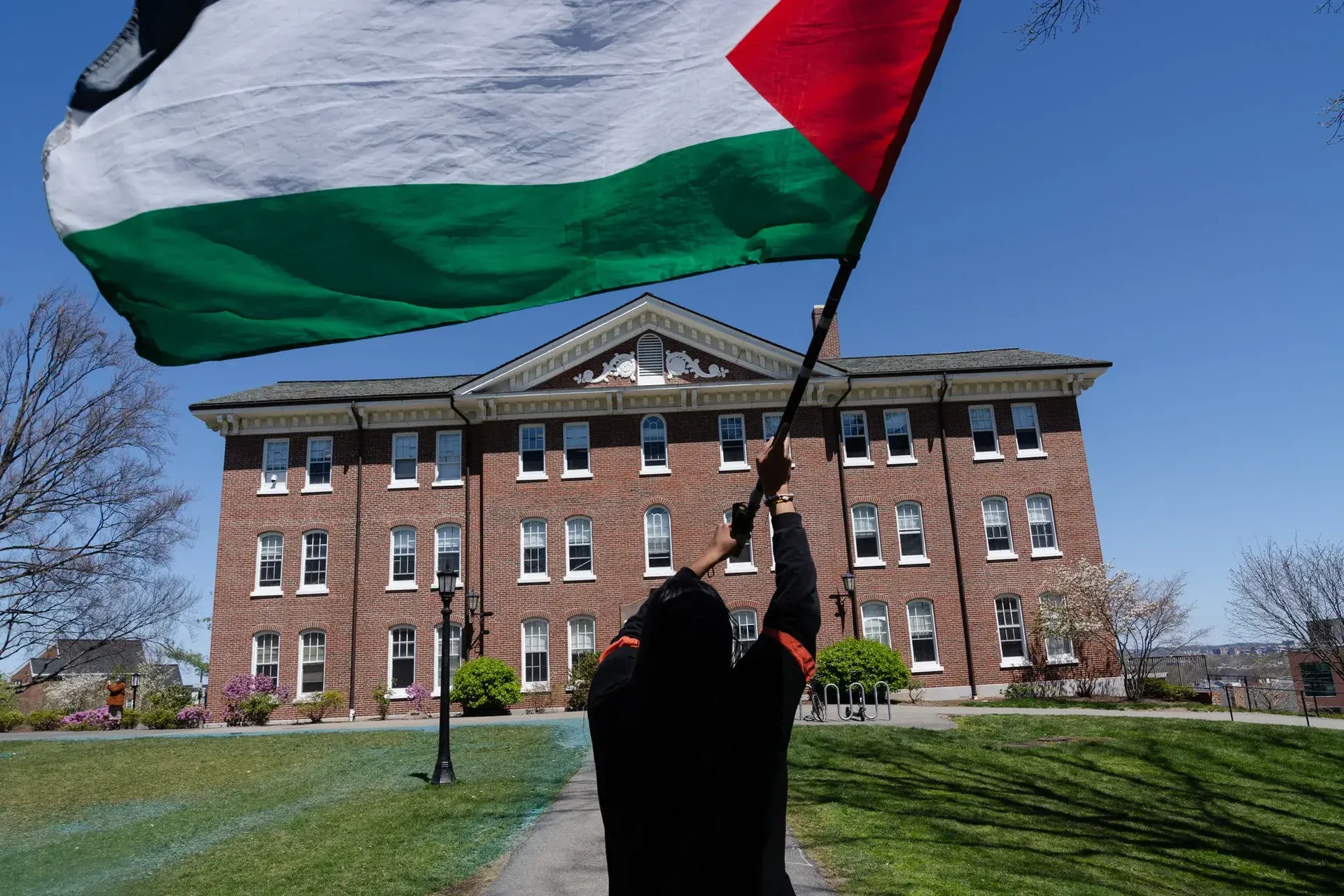 Someone waiving a massive Palestinian flag in front of a building with patches of grass on both sides, likely a university campus.