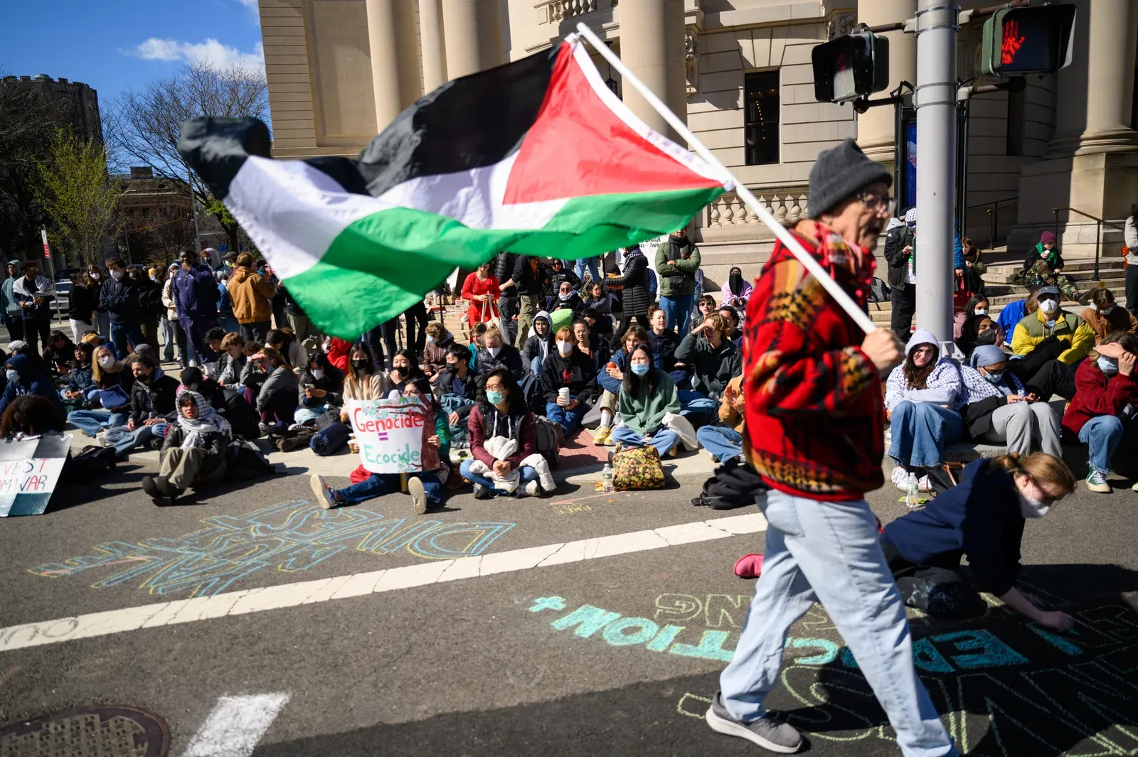 Dozens of people sitting with signs reading signs like "genocide = ecocide" and a man walking across with a large Palestinian flag.