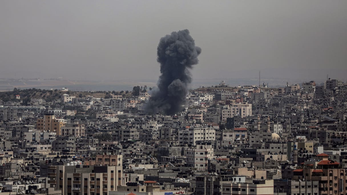 A dense city with a pillar of black smoke in the middle, rising from a probable airstrike